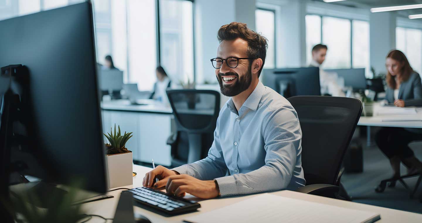 Lächelnder Mann mit Brille im Büro sitzt am Schreibtisch vor einem Monitor
