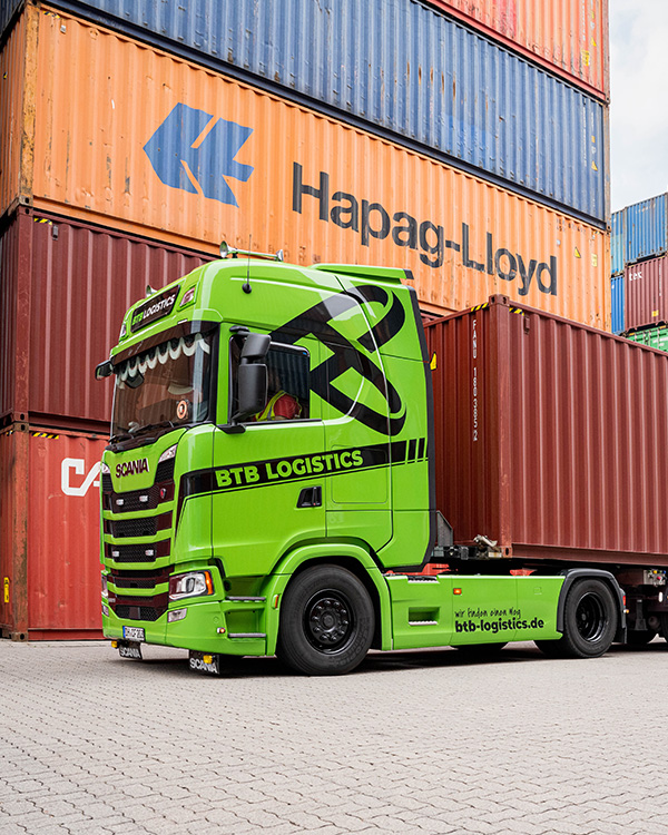 Grüner LKW von BTB beladen mit einem Container steht vor einem hohen Containerstapel